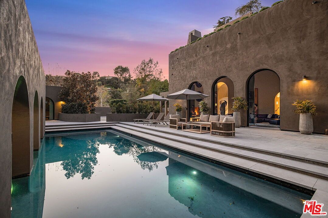 This sophisticated outdoor living space features a rectangular swimming pool set against a modern, earth-toned structure with arched doorways. The patio area is furnished with contemporary lounge seating and umbrellas, creating a serene environment for relaxation. The scene is captured at dusk, with the warm glow of interior lighting contrasting against the soft purple and orange hues of the twilight sky.