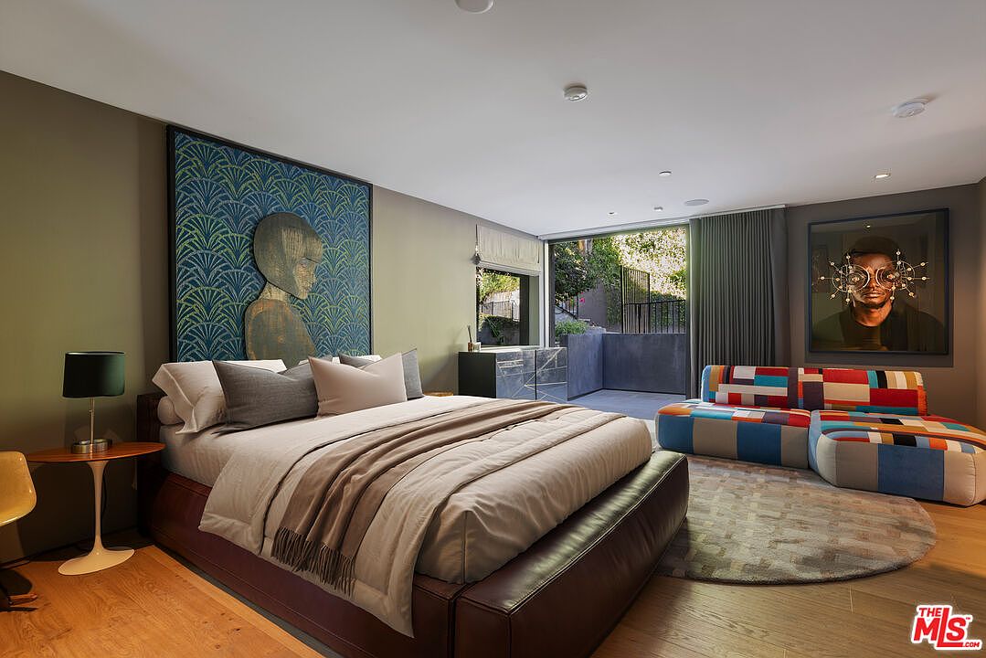 This contemporary primary bedroom features a low-profile bed with a dark leather frame, accented by a large, artistic wall panel with a geometric pattern. To the right, a vibrant, multi-colored modular sofa sits on a circular rug, while a large window provides a seamless transition to an outdoor patio area. The room is finished with warm wood flooring and modern art, creating a sophisticated and artistic living space.