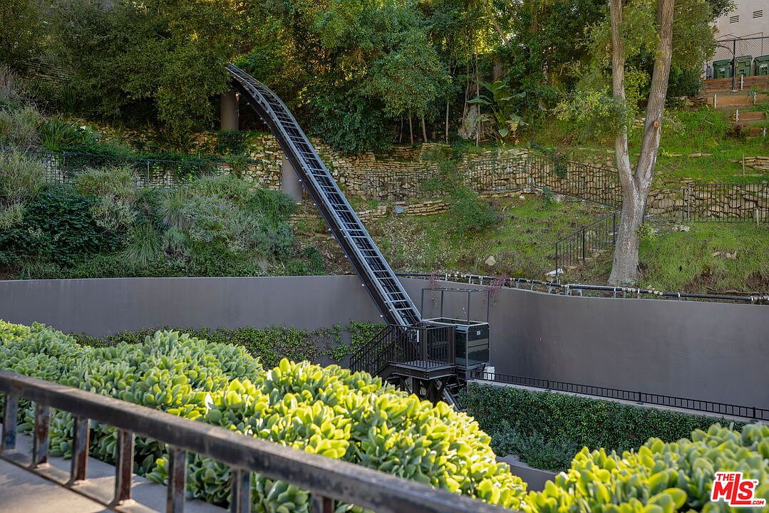 This exterior view highlights a steep, hillside property featuring a private funicular or inclined lift system designed to navigate the significant elevation change. The landscape includes lush green foliage, a stone retaining wall, and a metal staircase winding up the slope. The perspective is captured from a slightly elevated vantage point, emphasizing the unique engineering and terraced layout of the outdoor space.