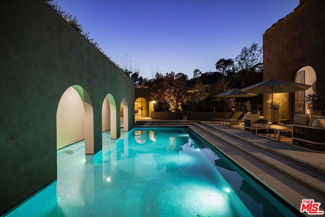 This elegant outdoor space features a rectangular swimming pool illuminated by soft underwater lighting, set against a Mediterranean-inspired structure with arched architectural details. A stone-paved patio area provides a comfortable lounge space with outdoor furniture and umbrellas, creating a serene atmosphere at dusk. The perspective captures the interplay between the glowing pool water and the warm ambient lighting of the surrounding landscape.