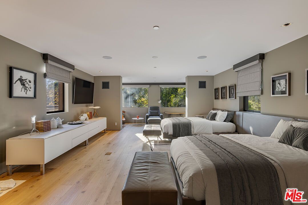 This spacious bedroom features two beds with neutral bedding and a long, low-profile white media console along the left wall. The room is finished with light wood flooring and soft gray walls, accented by framed artwork and large windows that provide natural light and views of the surrounding trees. A leather bench sits at the foot of the foreground bed, creating a comfortable and contemporary atmosphere.