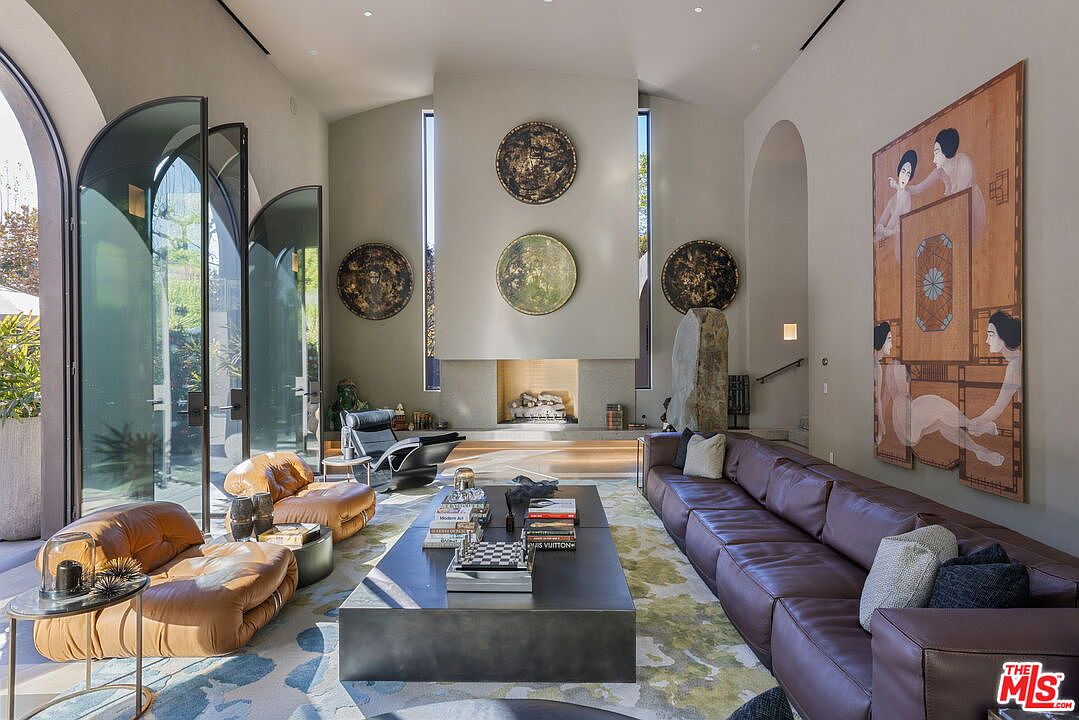 This expansive living room features a high, vaulted ceiling and a prominent central fireplace with a tall, light-toned surround. The space is furnished with a large, dark leather sectional, two low-profile tan leather lounge chairs, and a sleek, dark rectangular coffee table. Large, arched glass doors on the left provide a seamless connection to the outdoors, while a striking, large-scale figurative painting adorns the right wall, creating a sophisticated and artistic atmosphere.