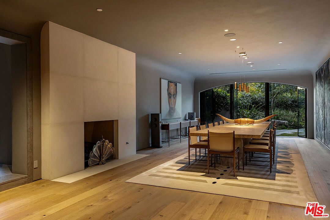 This sophisticated dining room features a long, light wood table centered on a large, patterned area rug, illuminated by a series of modern pendant lights. To the left, a tall, minimalist fireplace surround serves as a striking architectural focal point, while the far wall features expansive glass doors that open to a lush garden view. The space is defined by its neutral color palette, clean lines, and a blend of contemporary art and organic decor, creating an atmosphere of refined elegance.
