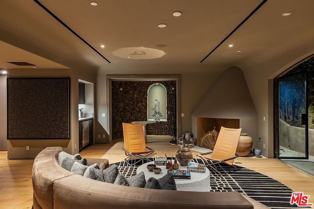 This sophisticated living room features a curved sofa facing a central coffee table and two leather lounge chairs, creating an inviting conversation area. A unique, sculptural fireplace with a smooth, tapered surround serves as a focal point, while a textured stone wall alcove in the background adds depth and visual interest. The space is finished with light wood flooring and a bold, geometric-patterned rug, offering a modern and artistic aesthetic.