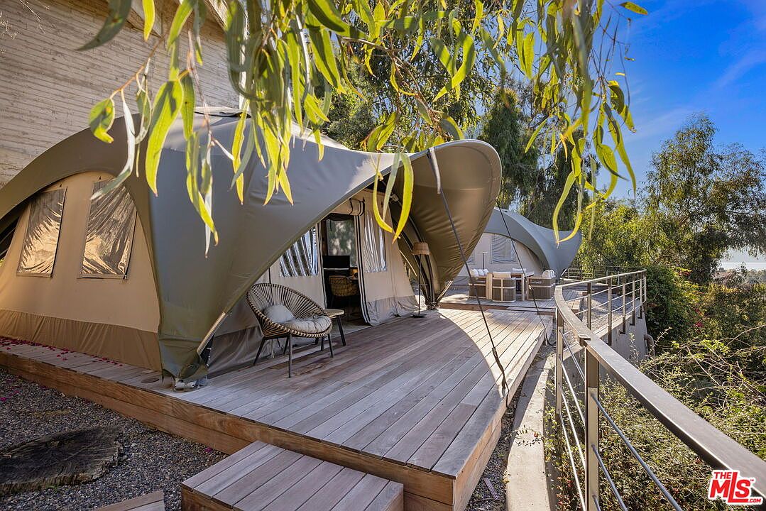 This image showcases a unique glamping-style setup on a spacious wooden deck, featuring two large, curved canvas tents. The foreground tent includes a modern woven chair on the deck, while the background tent is furnished with outdoor seating, all set against a backdrop of lush trees and a clear blue sky. The perspective is a slightly elevated eye-level shot, emphasizing the seamless blend of luxury camping and outdoor living space.