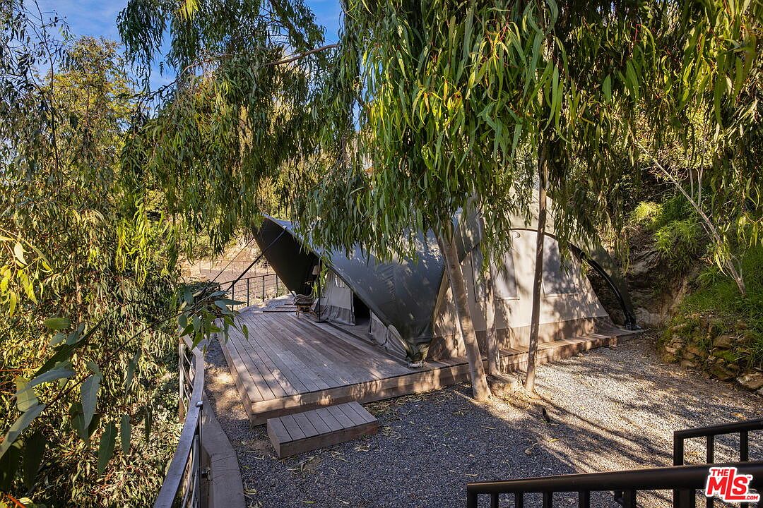 This unique outdoor living space features a luxury canvas tent structure set upon a raised wooden deck, nestled among mature eucalyptus trees. The scene captures a serene, glamping-style retreat with a gravel ground area and a small wooden step leading up to the deck. The perspective is from a slightly elevated vantage point, looking down through the foliage toward the secluded, nature-integrated dwelling.