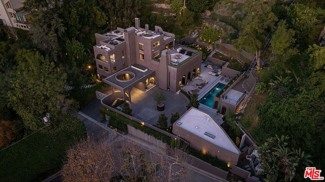 This high-angle aerial view captures a unique, multi-level contemporary residence nestled within a lush, wooded hillside. The property features a distinctive dark-toned exterior, a private swimming pool, multiple terraced outdoor lounge areas, and a gated motor court with a circular architectural cutout. The surrounding dense foliage and mature trees provide a secluded, tranquil atmosphere for this modern estate.