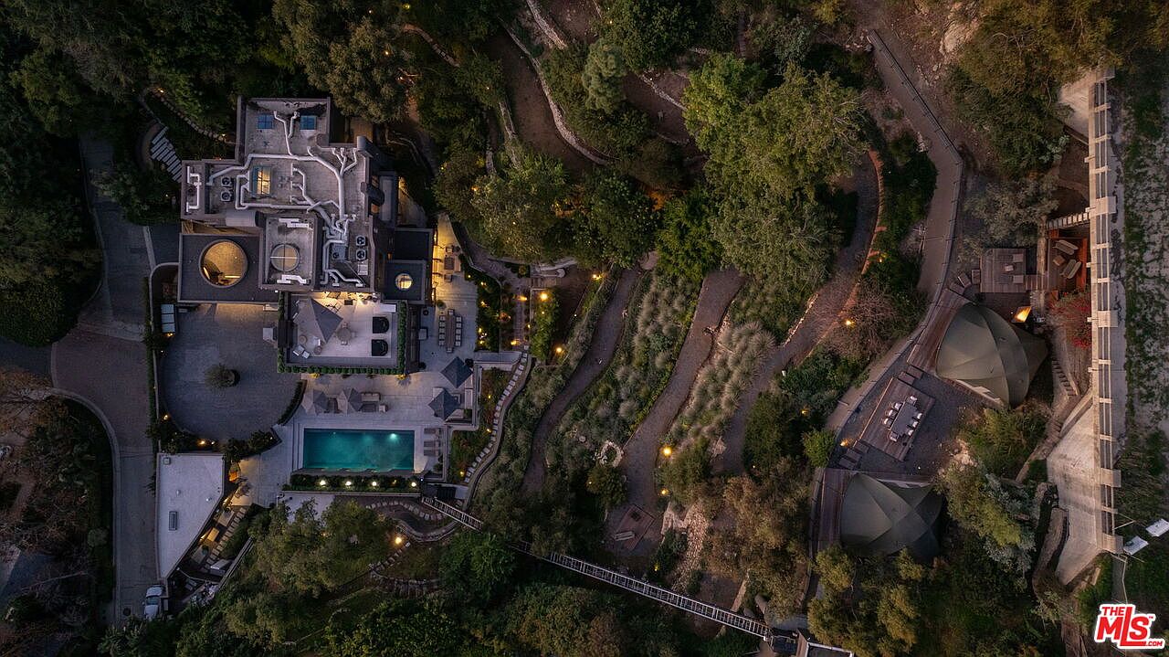 This high-angle aerial view captures a sprawling, multi-level estate nestled within a lush, wooded landscape. The property features a rectangular swimming pool, expansive stone-paved patio areas, and a unique bridge connecting different sections of the grounds. The architecture is modern and complex, with a flat-roof design and a network of outdoor pathways winding through the surrounding trees and garden features.
