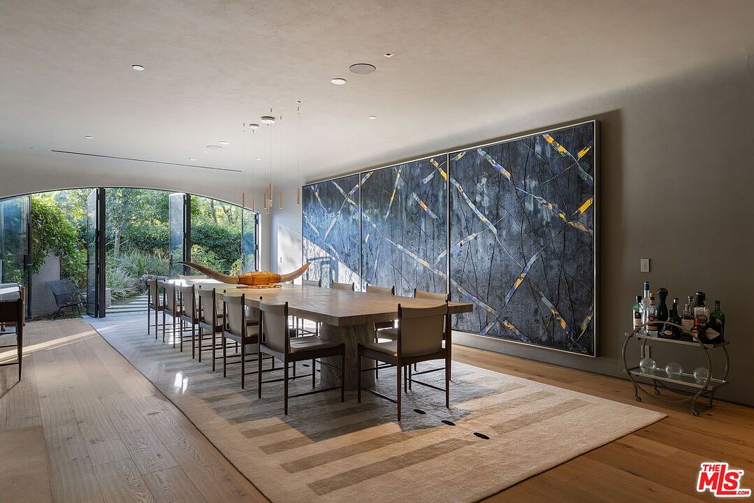 This sophisticated dining room features a long, light-toned wood table surrounded by modern chairs, set atop a large, textured area rug. A massive, multi-panel abstract artwork in shades of blue and gold dominates the wall, while a set of arched glass doors opens to a lush outdoor garden. The space is illuminated by a series of slender, minimalist pendant lights, creating an elegant and airy atmosphere.