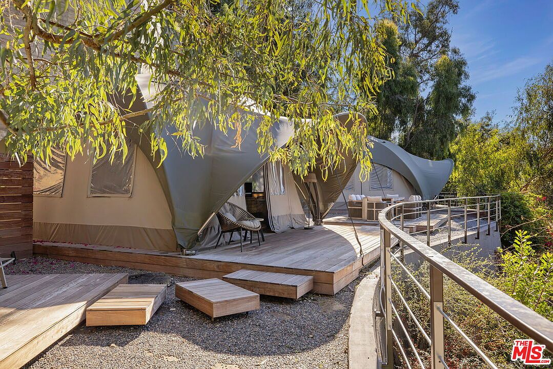 This image showcases a unique glamping-style structure featuring canvas-covered tent units set upon an expansive wooden deck. The outdoor living space is framed by a curved metal railing and surrounded by lush trees, creating a serene, nature-focused retreat. The perspective captures the transition from the gravel ground level up to the elevated deck, emphasizing the seamless integration of the structure with its natural environment.