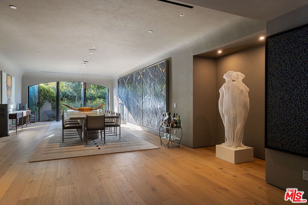 This sophisticated dining room features light wood flooring and a long, minimalist dining table centered on a large area rug. The space is defined by a striking, tall white sculpture in a recessed alcove and a massive, multi-panel abstract artwork spanning the side wall. Large, arched glass doors at the end of the room provide a seamless transition to the outdoor greenery, creating an airy and gallery-like atmosphere.
