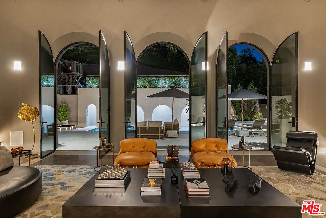 This sophisticated living room features a pair of cognac-colored leather armchairs facing a large, dark-toned coffee table, set against a backdrop of three expansive arched glass doors that open onto a pool area. The room is decorated with a patterned area rug, a gold-toned sculptural floor lamp, and modern lighting fixtures, creating a luxurious and inviting atmosphere. The seamless transition between the indoor seating area and the illuminated outdoor patio highlights the property's focus on indoor-outdoor living.