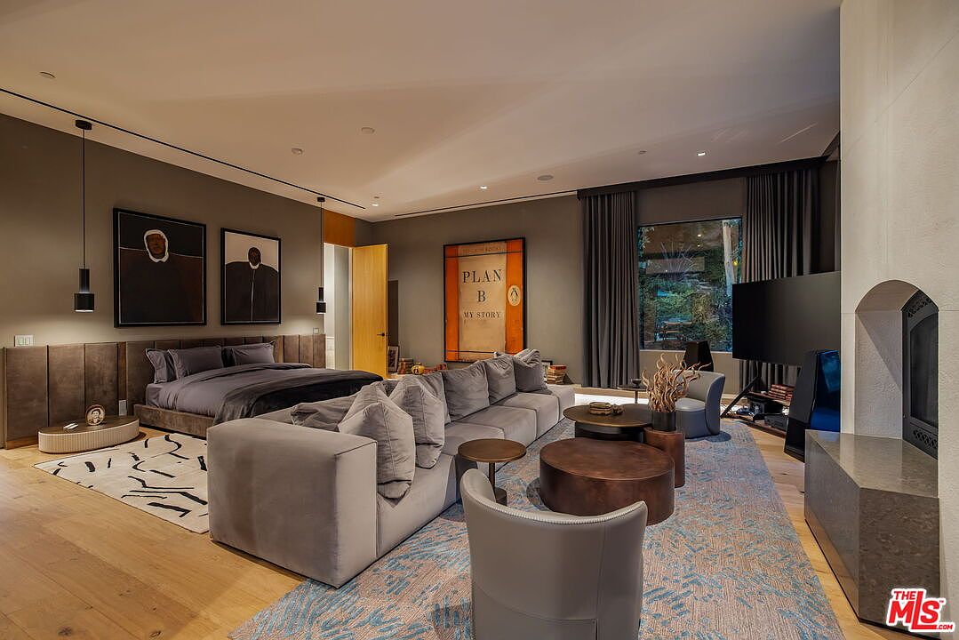 This expansive primary bedroom suite features a sophisticated, contemporary design with a neutral color palette and a large, plush sectional sofa positioned in the center of the room. The space includes a low-profile bed with a dark upholstered headboard, a prominent art piece titled 'Plan B', and a stone-clad chimney breast near a seating area. Large windows with dark drapery allow natural light to filter in, creating a luxurious and comfortable atmosphere.