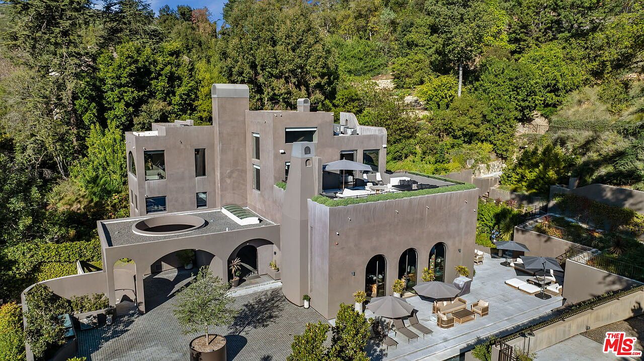 This aerial perspective captures a multi-level, contemporary residence with a distinctive stucco facade and a series of interconnected terraces. The property features a spacious paved courtyard, a rooftop deck with lounge seating, and a lower-level patio area furnished with umbrellas and outdoor furniture. Nestled against a lush, tree-covered hillside, the home showcases a unique architectural design with arched openings and a clean, modern aesthetic.