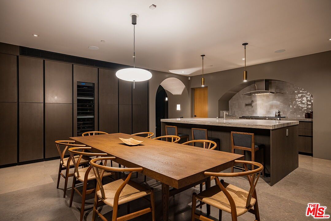 This sophisticated open-concept kitchen and dining area features a large wooden dining table surrounded by woven-seat chairs, positioned in front of a sleek kitchen island with stone countertops. The space is defined by dark, minimalist cabinetry, a built-in wine refrigerator, and a recessed cooking area with a stainless steel range hood. Warm pendant lighting hangs above both the dining table and the island, creating an inviting and modern atmosphere.