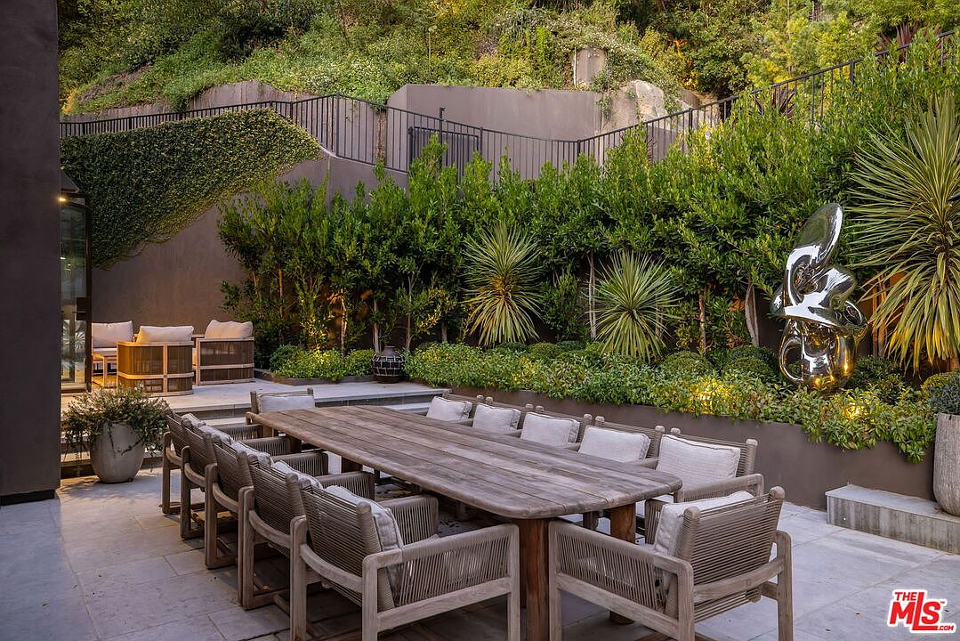 This sophisticated outdoor dining area features a long, light-toned wood table surrounded by matching chairs with woven detailing and neutral cushions. The patio is paved with large stone tiles and is bordered by a lush, manicured hedge and a modern abstract metal sculpture. In the background, a raised seating area with plush furniture provides a secondary space for relaxation, all set against a tiered hillside landscape.