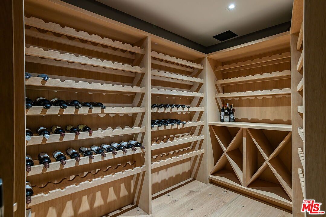 This custom-built wine cellar features extensive light wood shelving designed for both individual bottle storage and bulk display. The space includes angled racks for optimal bottle presentation and lower X-shaped bins for larger collections, all illuminated by recessed ceiling lighting. The perspective is a wide-angle view from the entrance, showcasing the organized and sophisticated storage capacity of the room.