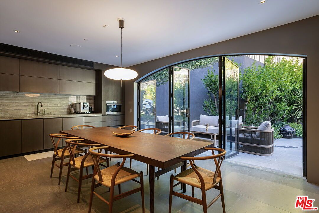 This contemporary dining area features a long, dark wood table surrounded by six light-toned wooden chairs with woven seats. To the left, a sleek, minimalist kitchen with dark cabinetry and a stone backsplash is visible, while a large, arched glass door system opens directly onto a private patio. The space is illuminated by a modern, low-hanging pendant light, creating a sophisticated and seamless transition between indoor dining and outdoor living.