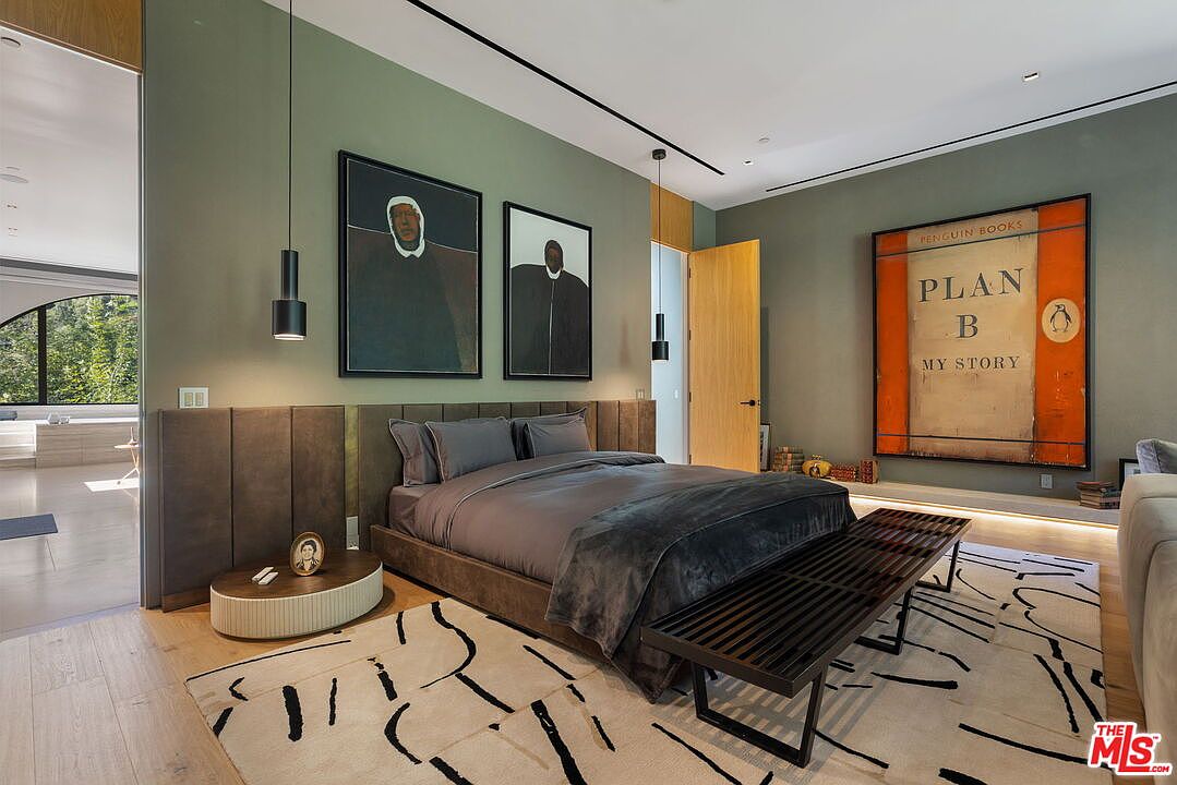 This sophisticated primary bedroom features a muted green accent wall behind a low-profile bed with a dark upholstered headboard. The room is decorated with two large abstract portraits and a prominent, oversized piece of wall art styled like a vintage book cover. A patterned area rug anchors the space, while a wooden bench sits at the foot of the bed, and a doorway leads to an adjacent bathroom area.