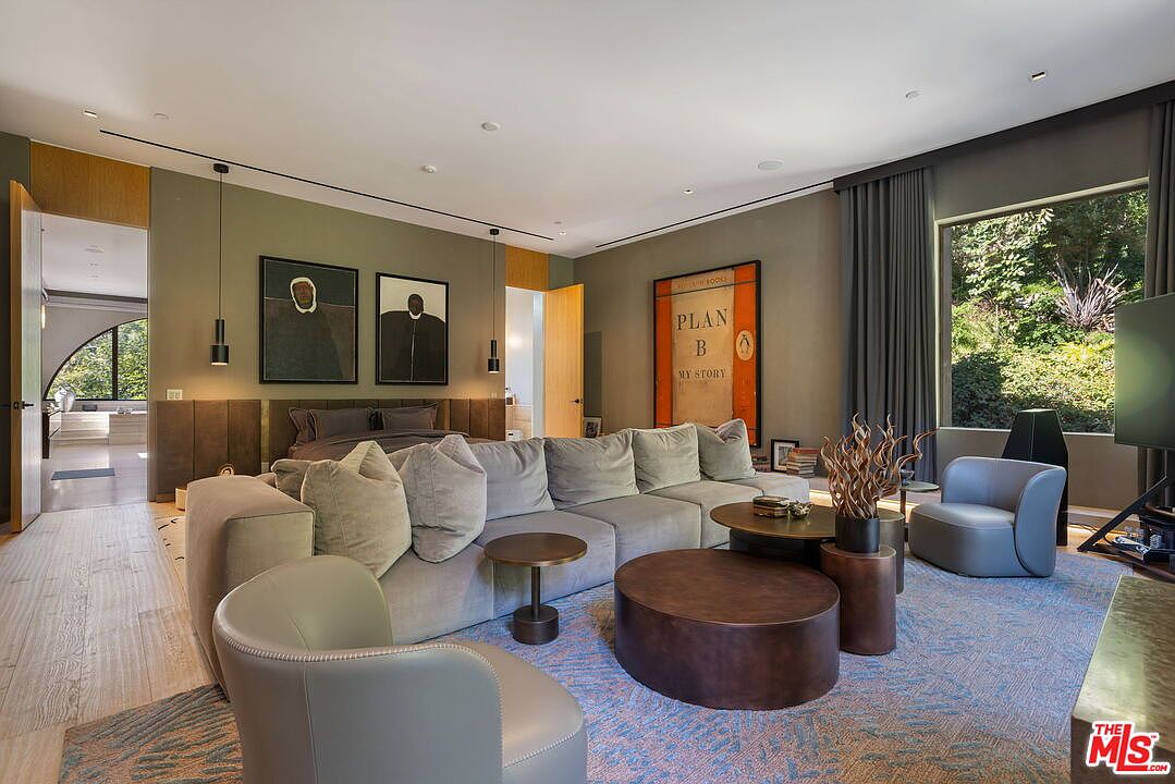 This spacious primary bedroom features a contemporary design with a large, plush sectional sofa positioned in the center of the room, creating a lounge area in front of the bed. The space is decorated with modern art, including a prominent 'Plan B' print, and is illuminated by sleek pendant lights and recessed lighting. Large windows provide natural light and views of the surrounding greenery, while a doorway leads to an adjacent bathroom, emphasizing an open and luxurious layout.