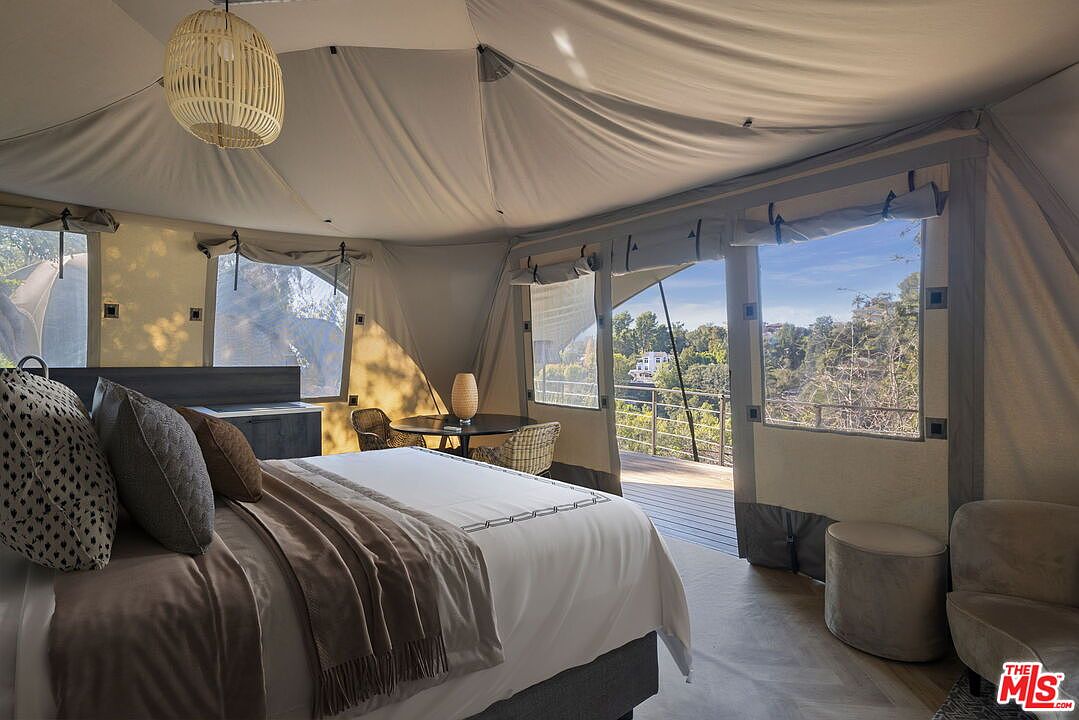This unique bedroom features a tent-like structure with soft, draped fabric walls and a high, vaulted ceiling. A plush bed with neutral-toned bedding sits in the foreground, while a small circular table and two woven chairs are positioned near large windows that open onto a wooden deck. The space offers a serene, glamping-inspired atmosphere with natural light streaming in to highlight the scenic outdoor view.