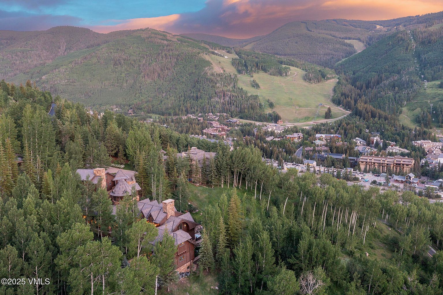 This high-angle aerial perspective captures a sprawling mountain estate nestled within a dense forest of aspen and pine trees. The multi-level home features a rustic architectural style with dark wood siding, complex rooflines, and multiple stone chimneys, blending seamlessly into the rugged landscape. In the background, a vast mountain range under a dramatic sunset sky provides a breathtaking backdrop to the surrounding valley and ski resort community.
