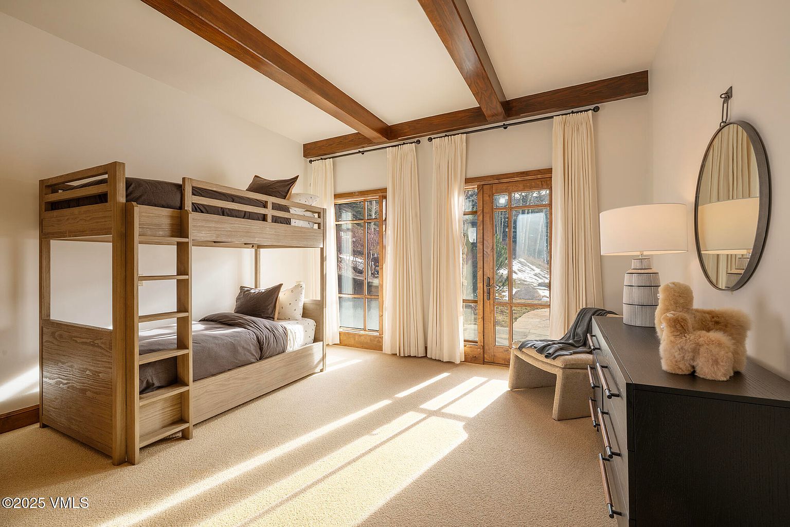 This bright and inviting guest bedroom features a light wood bunk bed set against a neutral wall, complemented by exposed wooden ceiling beams. Large glass doors with floor-to-ceiling curtains allow natural light to flood the space, highlighting the carpeted flooring and a dark wood dresser topped with a lamp and decorative accents. The room offers a cozy, rustic-inspired aesthetic with a view of the outdoor landscape through the glass.