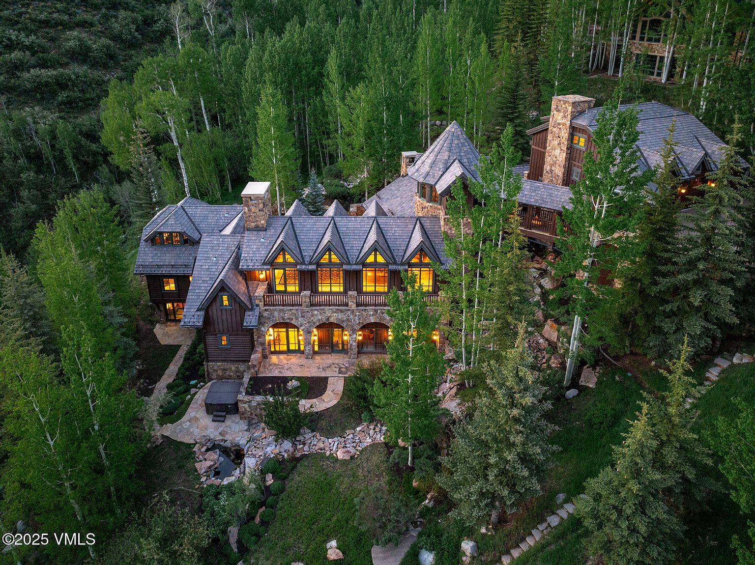 This high-angle aerial view captures a sprawling, multi-story mountain estate nestled within a dense forest of aspen and pine trees. The home features a complex roofline with multiple gables, stone and wood siding, and a warm, inviting glow emanating from numerous windows. A stone patio area with a hot tub and a small water feature sits at the base of the structure, emphasizing the property's seamless integration into its natural, secluded landscape.