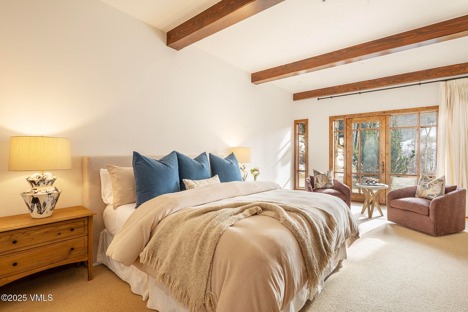 This serene primary bedroom features a neutral color palette with warm wood ceiling beams and soft carpeting. A plush bed with blue accent pillows serves as the focal point, flanked by light wood nightstands and patterned table lamps. Large glass doors provide access to the outdoors, allowing natural light to fill the space and offering a view of the snowy landscape.