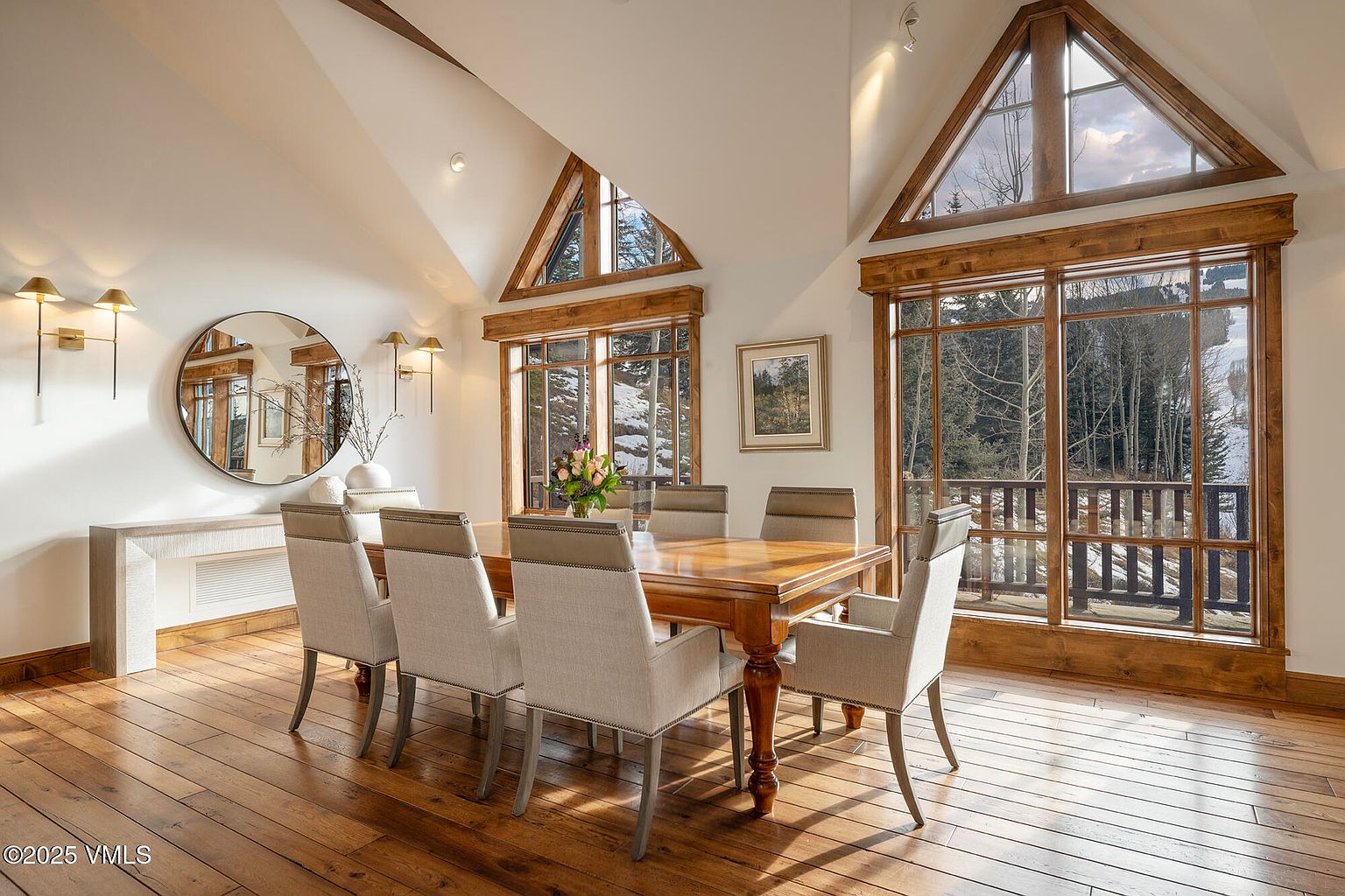 This elegant dining room features a large wooden table surrounded by six upholstered chairs, set against a backdrop of warm wood flooring and bright, neutral walls. The space is defined by its dramatic vaulted ceiling and expansive windows that frame a scenic, wooded outdoor view. A large circular mirror and wall-mounted light fixtures add a sophisticated touch to this bright, airy gathering space.