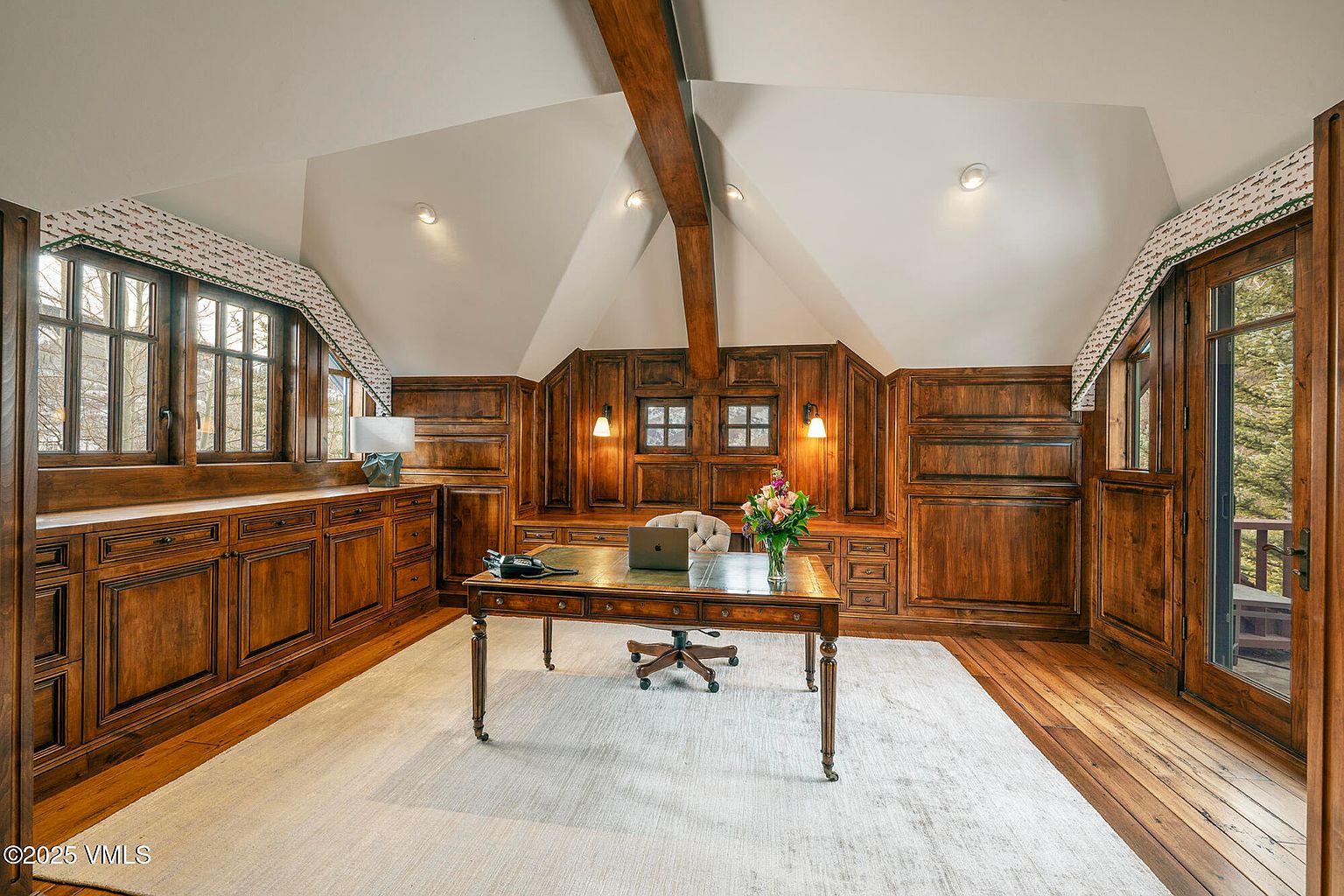 This sophisticated home office features rich, dark wood paneling throughout the walls and built-in cabinetry, creating a warm and professional atmosphere. A central wooden desk sits atop a light-colored area rug, complemented by a tufted office chair and a vase of fresh flowers. The room is illuminated by recessed lighting and wall-mounted sconces, with large windows and a glass-paneled door providing natural light and views of the surrounding trees.