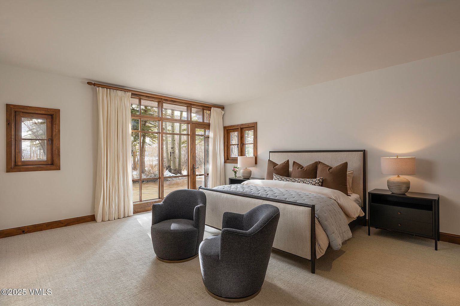 This serene primary bedroom features a neutral color palette, a plush upholstered bed, and two modern charcoal-colored swivel chairs positioned to enjoy the view. Large windows with floor-to-ceiling curtains frame a peaceful outdoor scene of trees and a stream, filling the room with natural light. The space is completed by two dark wood nightstands with matching lamps, creating a sophisticated and tranquil retreat.
