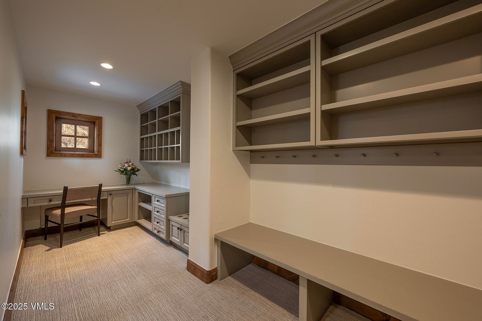 This functional home office or study area features built-in cabinetry, including a desk workspace and extensive shelving for storage. The room is finished with neutral-toned walls, carpeted flooring, and a wooden-framed window that provides natural light. A built-in bench with storage cubbies underneath adds utility to the space, creating a well-organized and versatile environment.