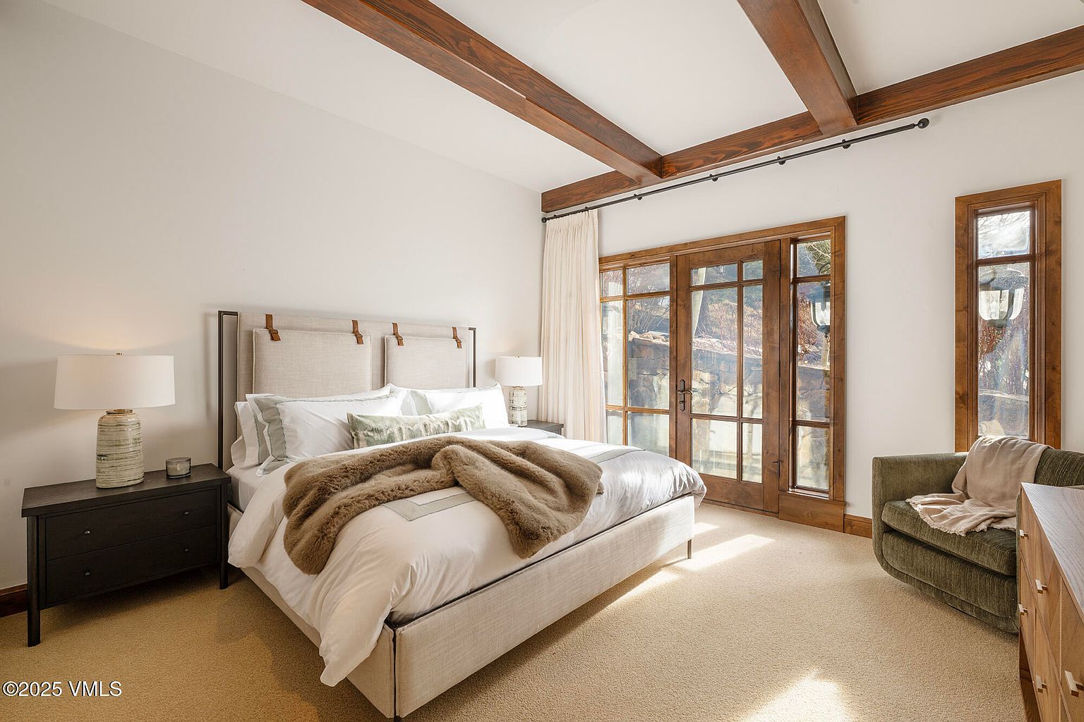This primary bedroom features a neutral color palette with a large upholstered headboard and a plush, textured throw blanket draped over the bed. The room is accented by exposed wooden ceiling beams and large glass doors that provide access to the outdoors, allowing for ample natural light. A comfortable green armchair sits in the corner, creating a cozy reading nook within the spacious, carpeted suite.