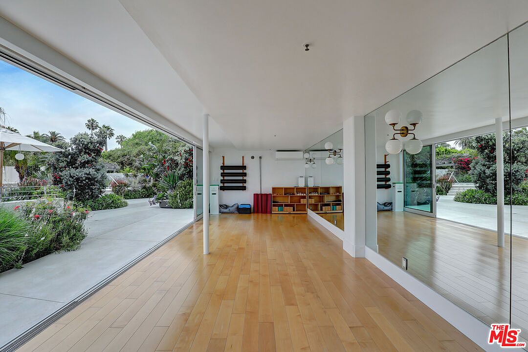 This bright, open-concept home gym features light wood flooring and expansive floor-to-ceiling glass sliding doors that seamlessly connect the indoor space to the outdoor patio. The room is equipped with wall-mounted exercise equipment, a wooden storage unit for fitness accessories, and a large wall mirror that enhances the sense of space. The minimalist design and direct access to the lush garden create a serene and inviting environment for wellness activities.