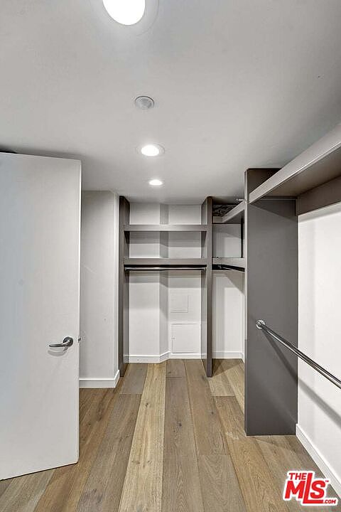 This walk-in closet features light wood-toned flooring and a clean, minimalist aesthetic with white walls and recessed ceiling lighting. The space is equipped with custom dark-toned shelving units and hanging rods, providing organized storage options. The perspective is from the doorway, looking into the closet to showcase its functional layout and modern design.