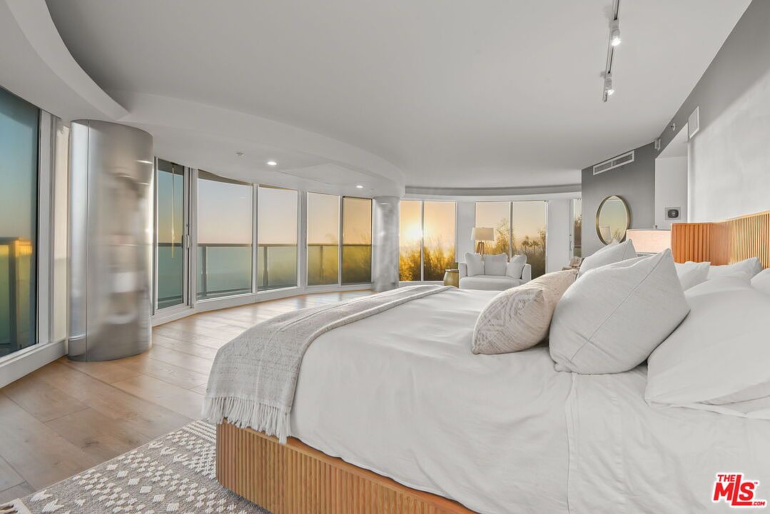 This spacious primary bedroom features a large bed with white linens and a light wood base, positioned to take advantage of the expansive, curved wall of windows. The room offers a panoramic view of the sunset over the ocean, creating a serene and luxurious atmosphere. A minimalist aesthetic is complemented by light wood flooring, a patterned area rug, and a small seating area near the glass.