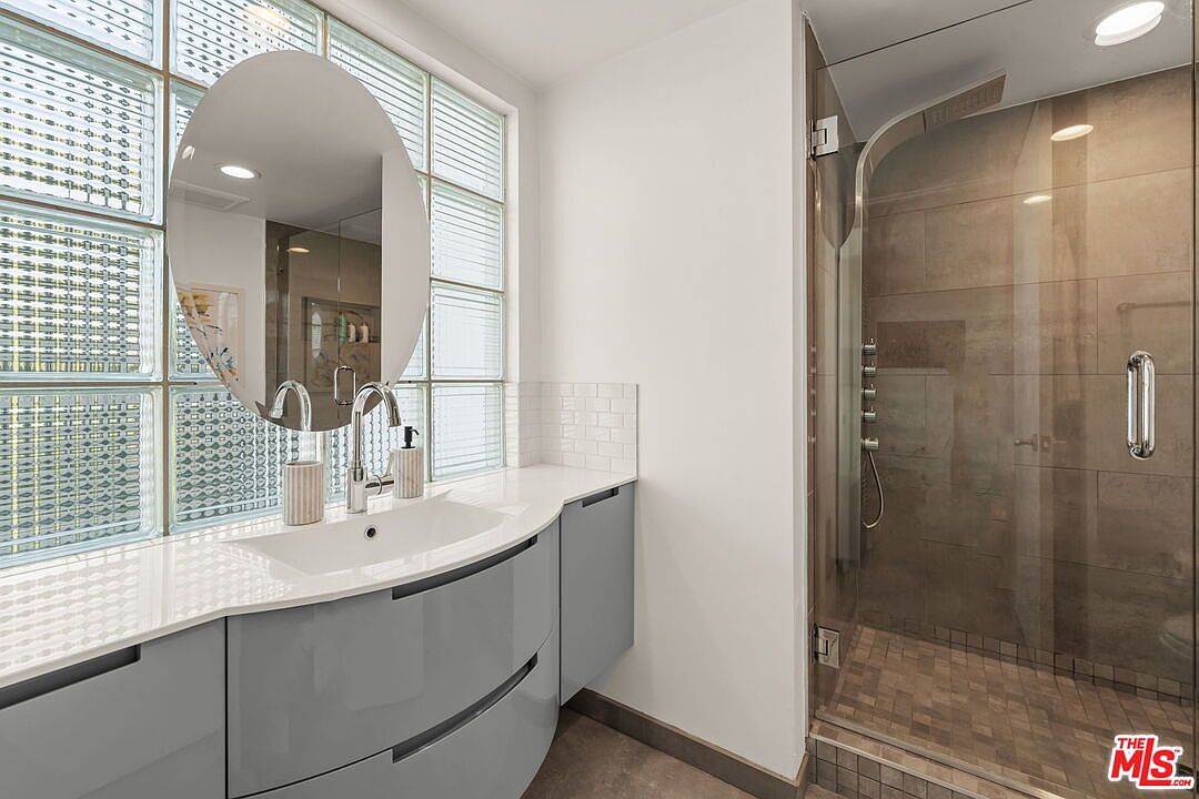 This modern bathroom features a sleek, curved vanity with a light-colored countertop and a unique oval mirror centered against a wall of glass block windows. To the right, a walk-in shower is enclosed by a glass door and finished with neutral-toned wall tiles and a textured floor. The perspective captures the clean lines and contemporary aesthetic of the space, emphasizing the contrast between the smooth vanity and the geometric glass blocks.