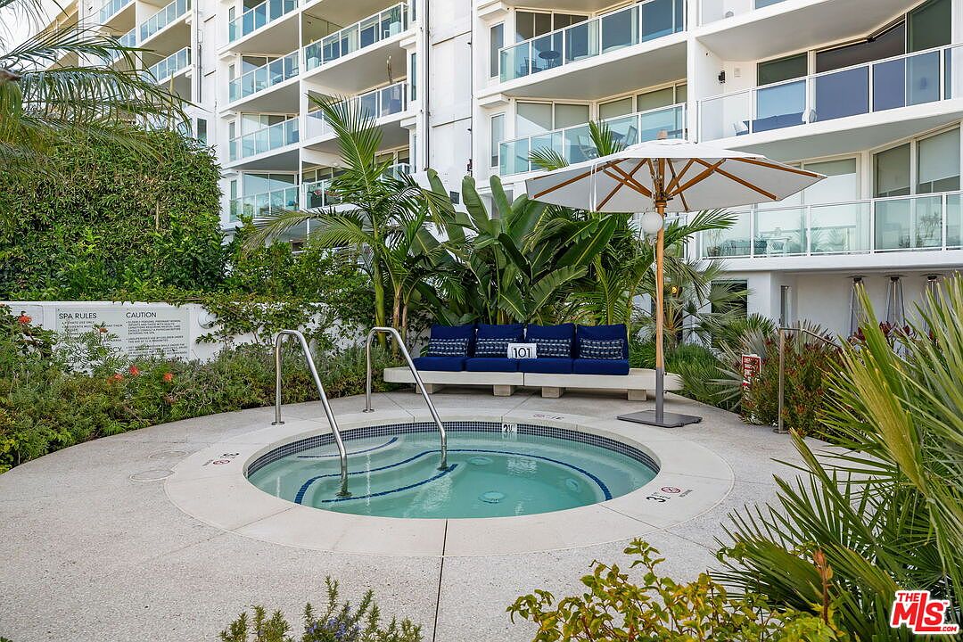 This image features a circular in-ground spa situated on a concrete patio, complete with metal handrails and integrated steps. Behind the spa, a long bench with blue cushions provides comfortable seating under a large white umbrella, surrounded by lush tropical landscaping. The scene is set against the backdrop of a multi-story residential building with glass-railed balconies, creating a resort-like atmosphere.