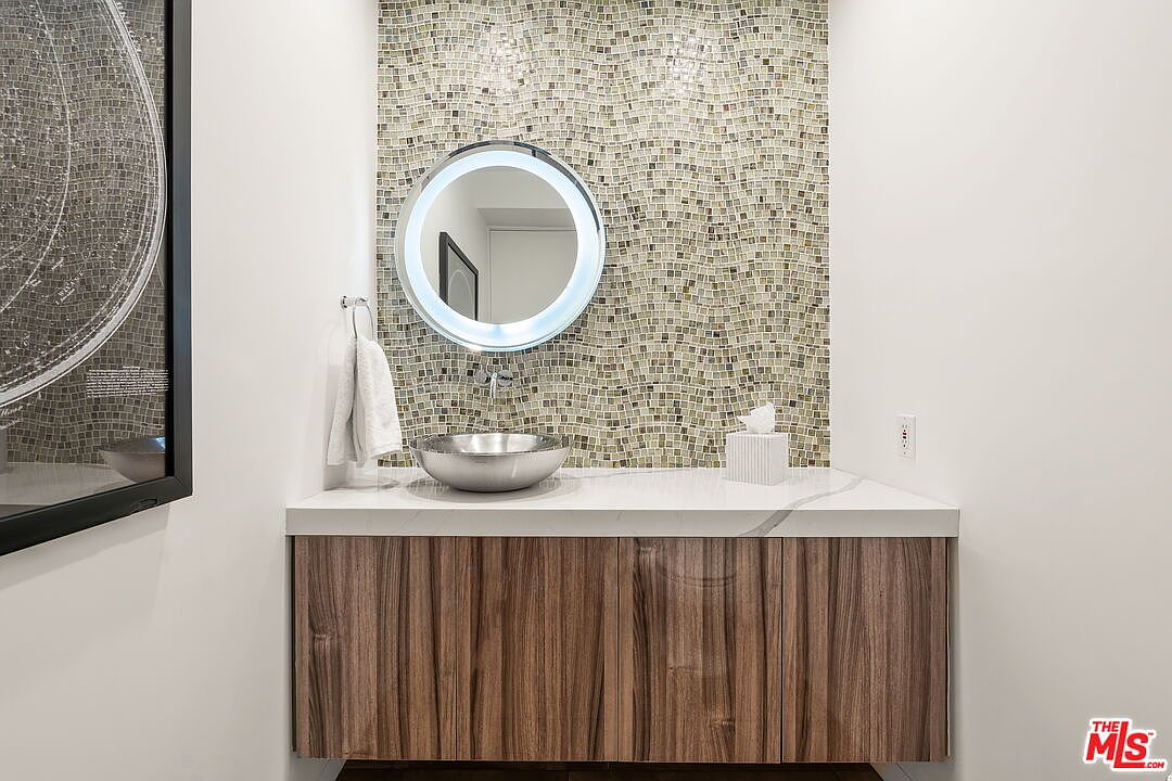 This modern powder room features a floating vanity with a wood-grain finish and a white countertop. A circular, backlit mirror is mounted against a decorative wall of small, textured mosaic tiles. A metallic vessel sink and a wall-mounted towel rack complete the contemporary aesthetic.