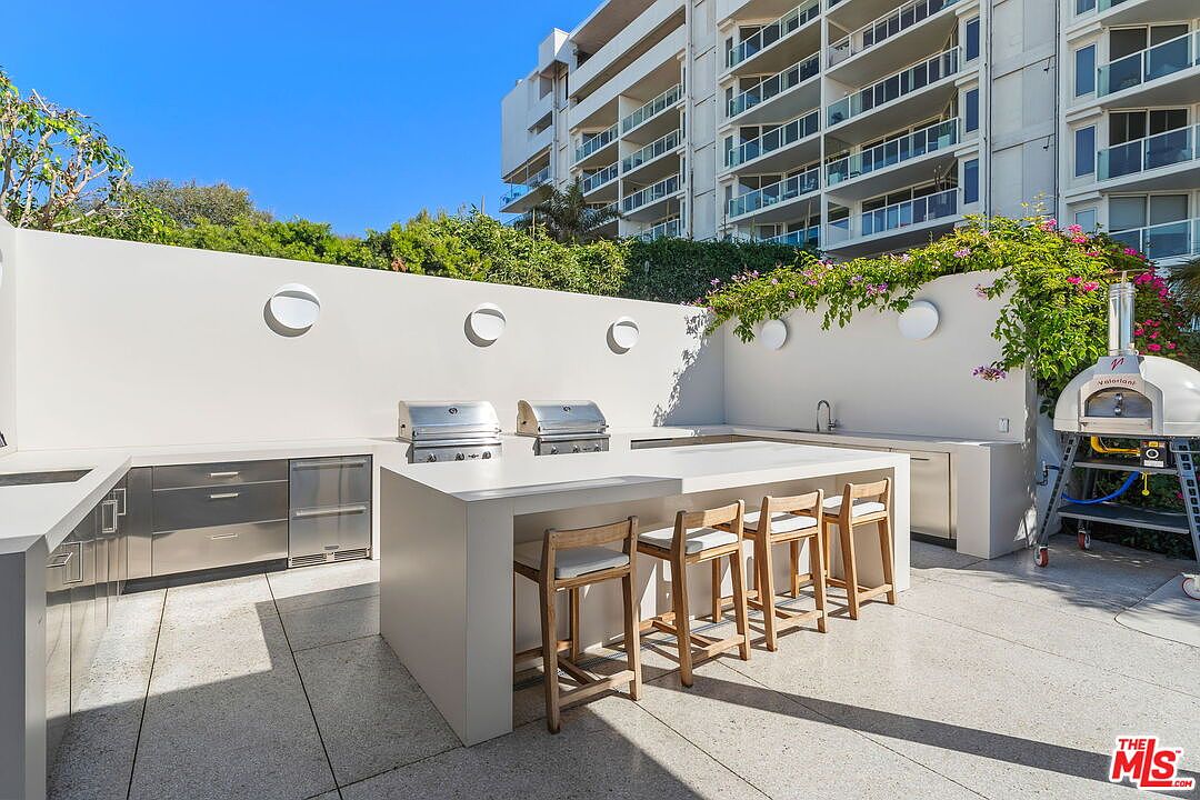 This outdoor kitchen and patio area features a sleek, minimalist design with a large central island, stainless steel built-in grills, and a portable pizza oven. Four light-wood bar stools are tucked under the island, which is set against a clean white wall adorned with circular wall sconces and climbing greenery. The space is paved with light-colored stone tiles, offering a bright and sophisticated setting for outdoor entertaining.