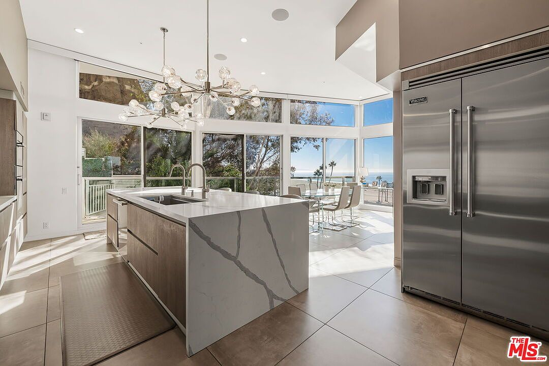This modern, open-concept kitchen features a large central island with a waterfall-edge stone countertop and light wood cabinetry. A striking, multi-armed chandelier hangs above the island, while a large stainless steel refrigerator stands to the right. The space is bathed in natural light from expansive windows that offer a clear view of the ocean and surrounding landscape.