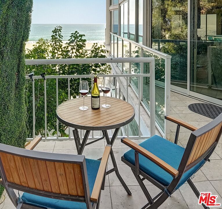 This balcony offers a serene, elevated view of the ocean and sandy beach, framed by lush green foliage. The space is furnished with a small, round wooden table and two chairs with blue cushions, creating an inviting spot for relaxation. Large glass sliding doors provide seamless access to the interior, enhancing the connection between the living space and the coastal scenery.