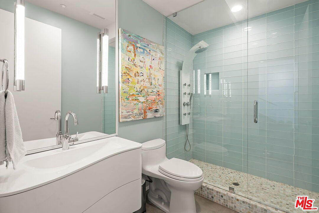 This contemporary bathroom features a sleek white vanity with a modern faucet and a wall-mounted mirror flanked by vertical light fixtures. The shower area is enclosed by a glass panel and showcases light blue subway-style wall tiles paired with a multi-colored mosaic tile floor. An abstract, colorful painting adds a vibrant artistic touch to the light-toned walls, creating a bright and clean aesthetic.