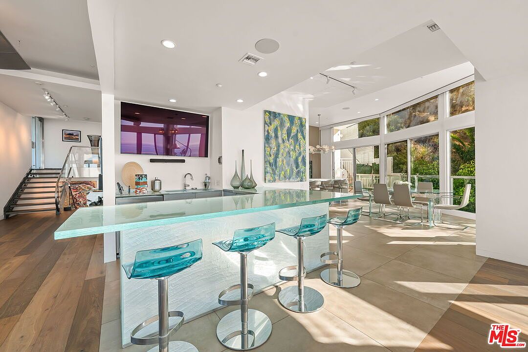This modern, open-concept kitchen and bar area features a prominent island with a thick, translucent glass countertop and three vibrant blue acrylic bar stools. The space is bright and airy, highlighted by large windows that provide a view of the outdoor landscape and a staircase leading to an upper level. The contemporary design is complemented by light-toned tile flooring and a large piece of abstract wall art, creating a sophisticated and inviting atmosphere.