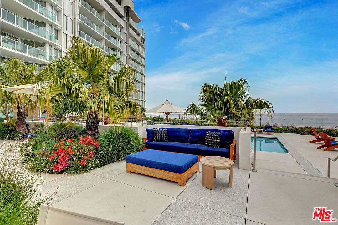 This expansive outdoor terrace features a comfortable seating area with a deep blue sofa and matching ottoman, set against a backdrop of lush palm trees and a modern multi-story building. To the side, a pristine swimming pool overlooks the ocean, creating a serene and luxurious atmosphere. The concrete patio provides a clean, contemporary space perfect for relaxation and taking in the coastal views.