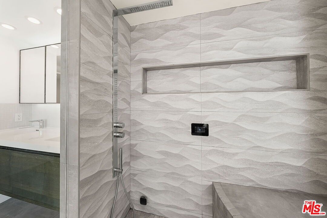 This modern bathroom features a walk-in shower with textured, wave-patterned wall tiles and a built-in recessed niche for storage. A sleek, wall-mounted shower fixture with multiple controls is positioned next to a glass partition that looks out toward a vanity area with a mirror and integrated lighting. The space conveys a contemporary, spa-like aesthetic with clean lines and a neutral color palette.