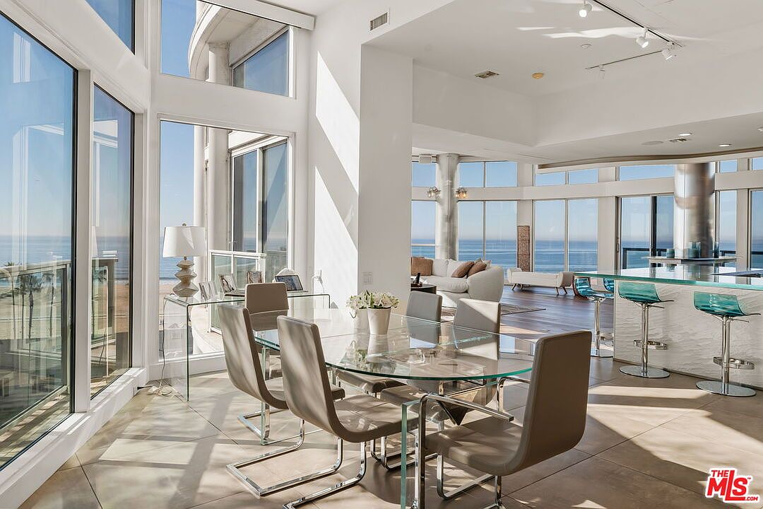 This bright and airy dining area features a glass-topped table surrounded by six modern, neutral-toned chairs. The space is defined by expansive floor-to-ceiling windows that offer panoramic ocean views and flood the room with natural light. In the background, a kitchen island with bar seating and a living area are visible, creating an open-concept layout that emphasizes a luxurious, coastal lifestyle.