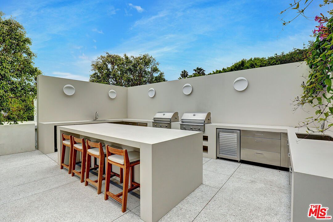 This modern outdoor kitchen area features a sleek, minimalist design with a large, light-toned stone island and integrated bar seating. The space is equipped with dual stainless steel grills, a built-in refrigerator, and a sink, all set against a clean, neutral-colored wall accented by circular wall-mounted lights. The perspective is a wide-angle shot capturing the functional layout of this sophisticated al fresco dining and cooking space.