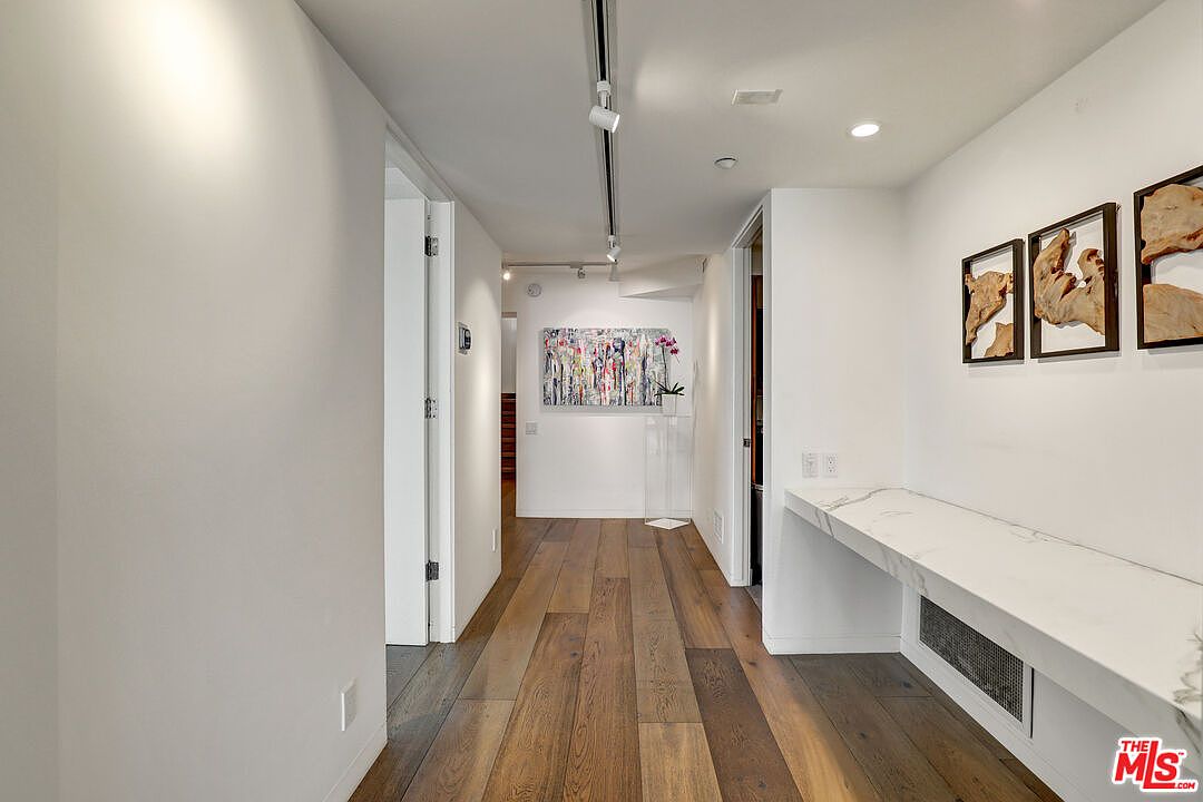 This bright, modern hallway features wide-plank wood flooring and crisp white walls, creating a clean and minimalist aesthetic. To the right, a long, sleek console with a white stone top provides a functional surface, complemented by three framed abstract art pieces on the wall. The perspective looks down the corridor toward a vibrant, colorful painting at the far end, with recessed lighting and track lighting overhead enhancing the gallery-like atmosphere.